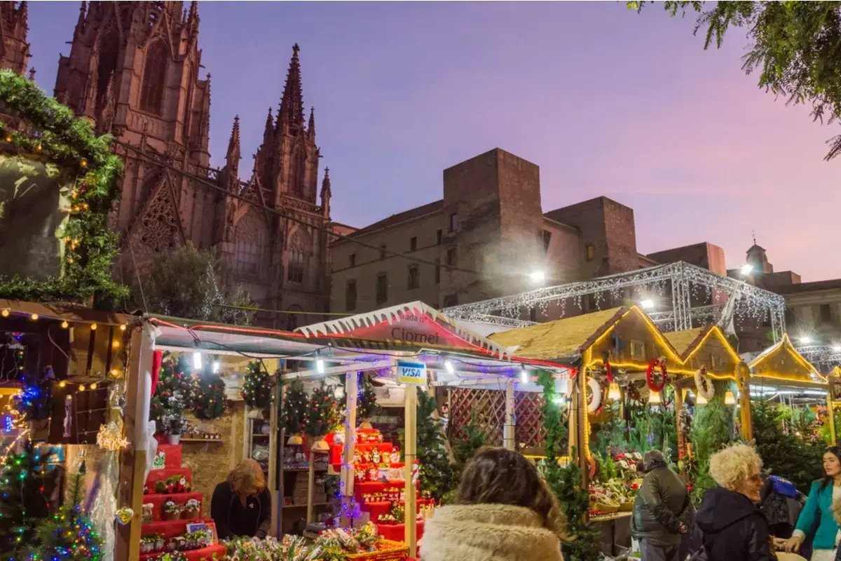 Christmas market at the Fira de Santa Llúcia in Barcelona, with festive lights, craft stalls, Christmas decorations and the illuminated Barcelona Cathedral in the background, with people walking and enjoying the warm holiday atmosphere.